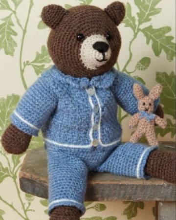 A crocheted bear wearing blue pajamas sits on a wooden bench in front of a leafy wallpapered wall. 
