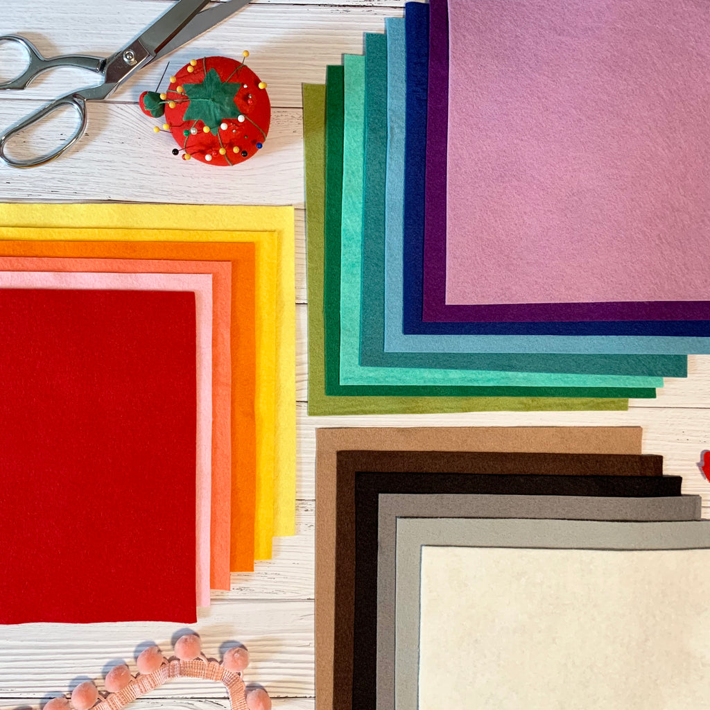 Flat lay with square sheets of felt in rainbow colors, a par of scissors, a tomato pin cushion, and a strand of pom pom trip