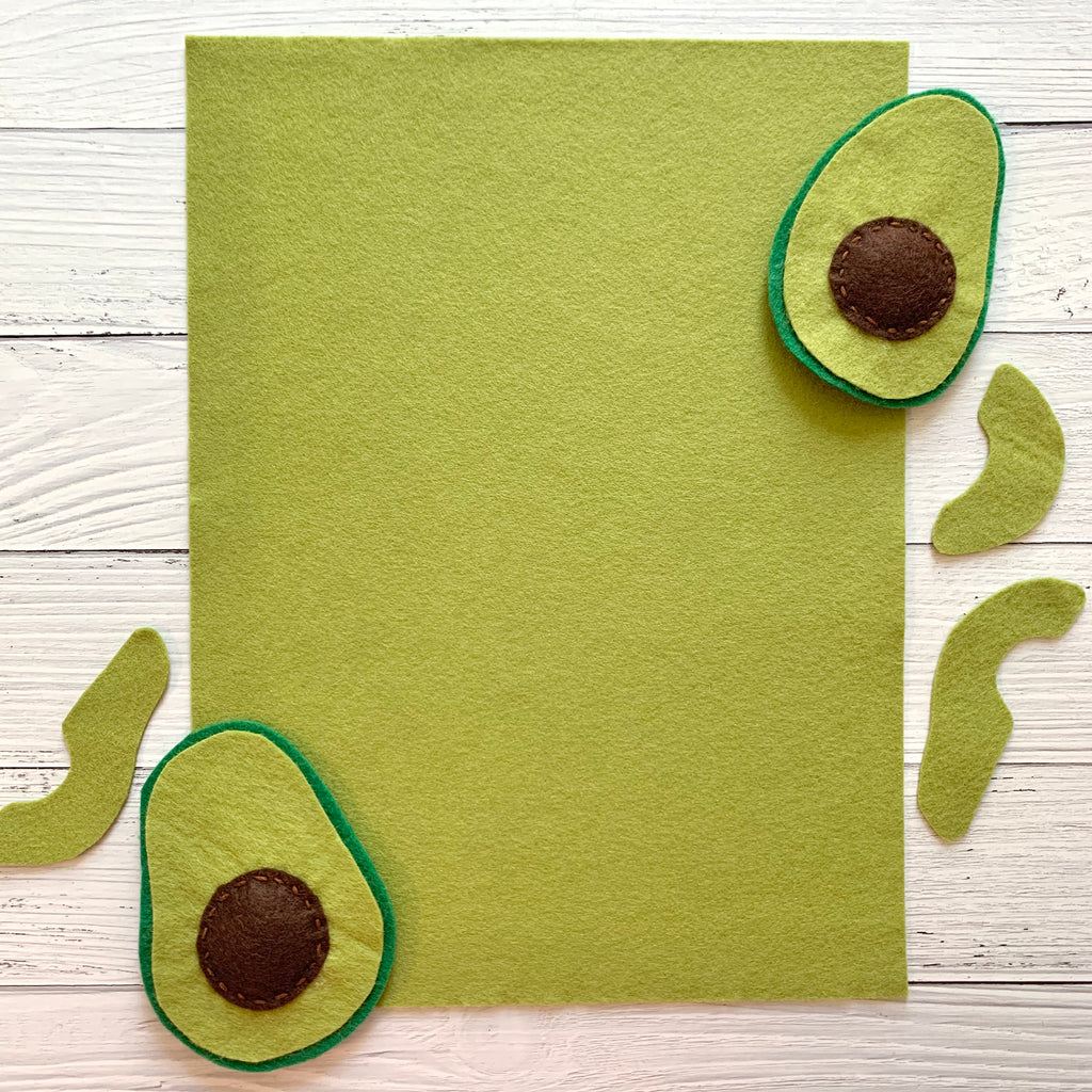 one 8 x 10 sheet of avocado green colored felt on a white wood background with cut out felt avocado shares on top