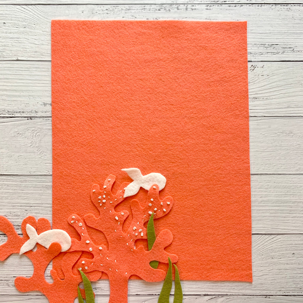 one 8 x 10 sheet of coral colored felt on a white wood background with coral cut out shapes, sea weed, and white fish cut out of felt.