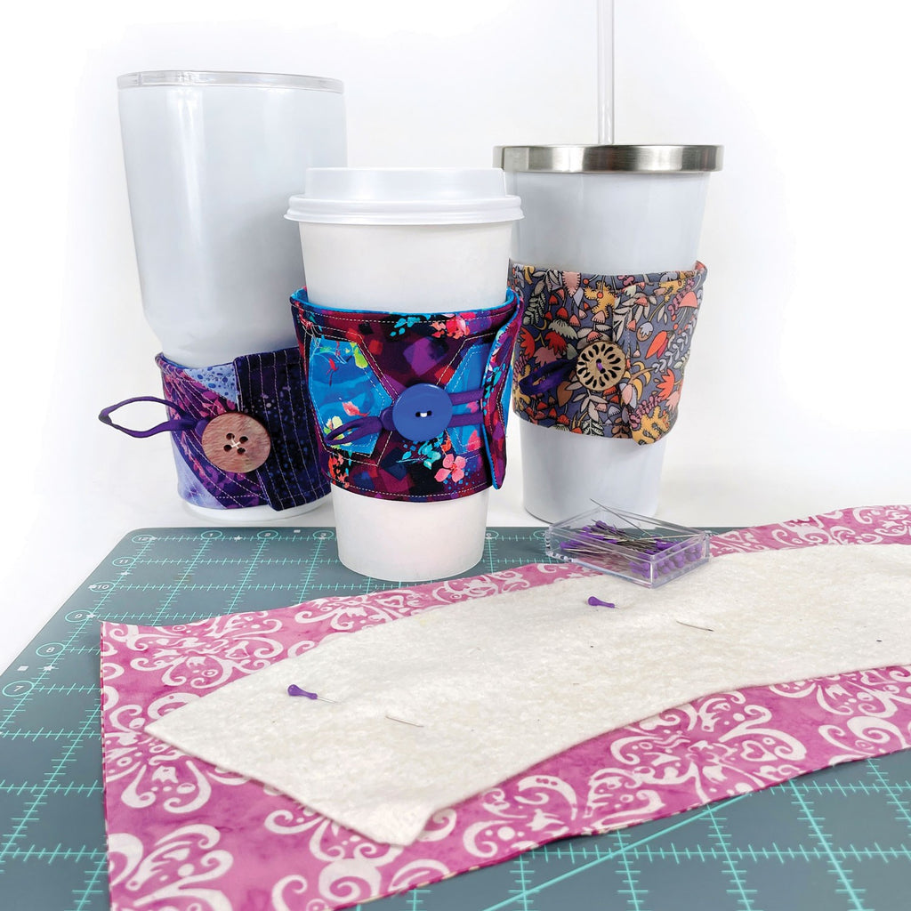 Three drink cups with drink cozies sit on a quilting cutting mat . 