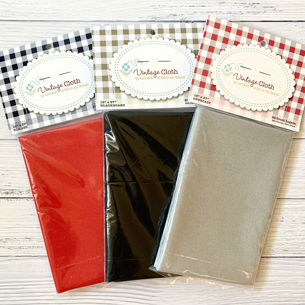 Three packages of 25 count cross stitch fabric on a white wood background. Packages are red, black, & gray.